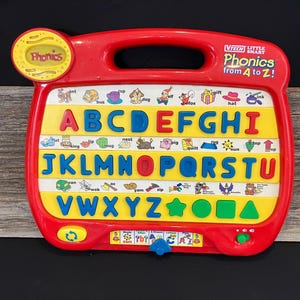May include: A red and yellow plastic toy learning tool with a handle. The toy features a colorful alphabet with pictures and words for each letter. The toy is labeled "VTech Little Smart Phonics from A to Z!"