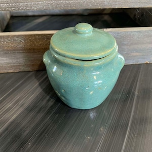 Può includere: Un piccolo barattolo di ceramica blu-verde con un coperchio. Il barattolo ha una superficie leggermente ruvida e una forma arrotondata.