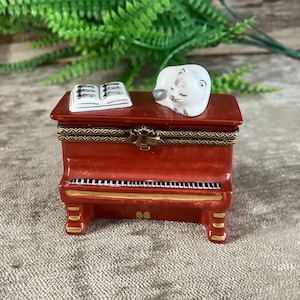 May include: A small, red, piano-shaped porcelain trinket box with a hinged lid. The top features a white, sleeping cat figurine and an open book with black musical notes. The piano has gold-painted accents and a decorative brass-coloured metal clasp.