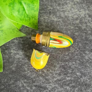 Puede incluir: Un vial de perfume vintage con una tapa de latón y un diseño colorido y arremolinado en amarillo, verde, rojo y blanco. El vial está abierto, revelando el interior. La tapa está separada y el vial está sobre un fondo gris.