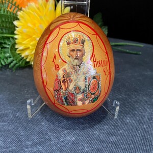 Peut inclure: Un œuf en bois peint à la main avec un fond doré et une bordure rouge. L'œuf représente Saint Nicolas, une icône religieuse, avec une couronne dorée et une robe rouge et dorée. Le texte "Никола" est écrit en écriture cyrillique sur l'œuf.