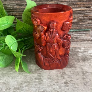 May include: A reddish-brown, carved wooden pen holder. The cylindrical holder features detailed relief carvings of a robed figure holding a scroll and basket. The pen holder is set against a backdrop of green leaves and a wooden surface.