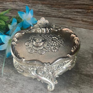 Puede incluir: Joyero plateado y ovalado con detalles florales. La tapa presenta un diseño floral en relieve, con detalles en los laterales y patas curvas. Flores azules artificiales se ven en el fondo. El joyero es de metal.