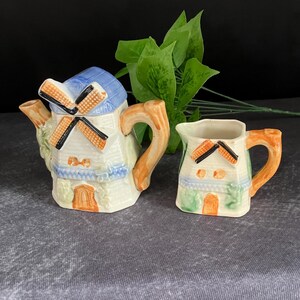 Può includere: Un set di teiera e lattiera in ceramica. La teiera ha la forma di un mulino a vento con tetto blu e pale arancioni. La lattiera è una casa in miniatura. Entrambi i pezzi hanno manici arancioni.