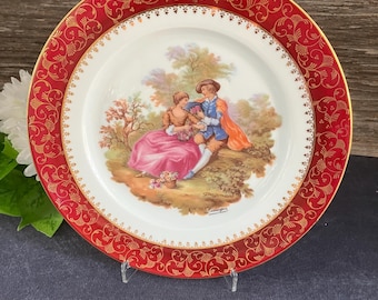 May include: A decorative porcelain plate with a red and gold ornate border. The center features a pastoral scene with two figures in period clothing. The plate is supported by a clear stand, showcasing the design.