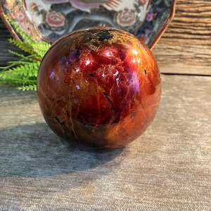 Könnte beinhalten: Eine dekorative Kugel mit glänzendem, rotbraunem Finish. Die Kugel hat ein marmoriertes Aussehen mit dunklen Adern und Glanzlichtern. Im Hintergrund befindet sich ein dekorativer Teller mit Vogel- und Blumenmotiven.