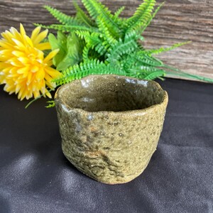 Könnte beinhalten: Eine grüne Teetasse aus Keramik mit einer rauen, strukturierten Oberfläche. Die Tasse hat einen breiten, abgerundeten Rand und einen leicht ausgestellten Boden.