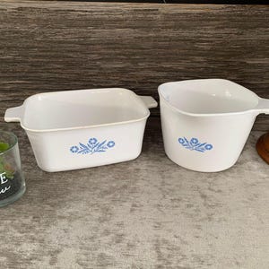 Puede incluir: Dos platos para hornear de cerámica blanca con diseños florales azules. Los platos están sobre una superficie de madera.