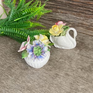May include: Two porcelain figurines of flowers in white vases. One vase is shaped like a swan and the other is round. The flowers are yellow, pink, and blue.