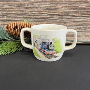 Könnte beinhalten: Eine weiße Keramiktasse mit zwei Henkeln zeigt ein Cartoon-Bild von Thomas der Lokomotive, der einen Zug durch einen Tunnel zieht. Die Tasse hat ein grünes, gelbes und blaues Design.