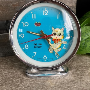 Op de afbeelding: Een vintage wekker met een chromen frame en een lichtblauwe wijzerplaat. De klok heeft een cartoon kat illustratie, een rode secondewijzer en het woord "HERO". De klok is ongeveer 10 cm hoog.