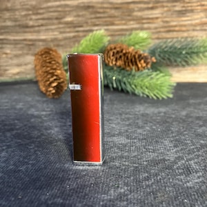Puede incluir: Un encendedor rojo y plateado. El encendedor tiene forma rectangular con una parte delantera roja y laterales plateados. El fondo incluye agujas de pino y piñas.
