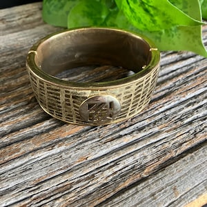May include: A gold-coloured metal cuff bracelet with a textured, grid-like pattern. The bracelet has an oval-shaped accent with an engraved design. The jewellery is displayed on a weathered wooden surface.