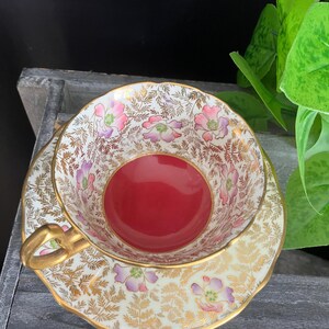 May include: A vintage teacup and saucer set. The cup has a red interior and is decorated with pink and purple floral patterns and gold accents. The saucer mirrors the cup's design, with a gold rim. The set is on a wooden surface.