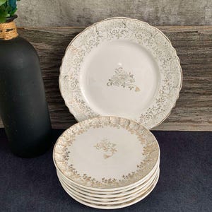 May include: A set of eight white ceramic dessert plates with a gold floral pattern and a gold rim. The plates are stacked on top of each other.