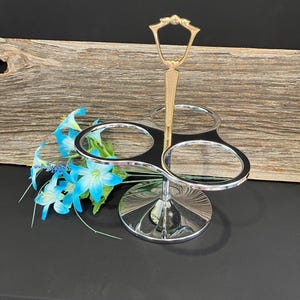May include: A silver and gold three-tiered condiment holder with a handle. The holder has three round glass bowls and a round base.