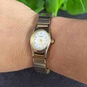 May include: A vintage wristwatch with a gold-tone bezel and a silver-tone band. The watch face is white with black numerals and the word "GITANOE". The watch is approximately 0.75 inches in diameter.