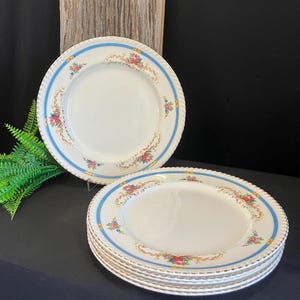 以下が含まれることがあります： 青い縁と花柄模様の白いセラミックプレートのセット。各プレートはスカラップエッジと金色のトリムが付いています。花柄のデザインには、ピンク、赤、黄色の花が含まれています。プレートは暗い表面に積み重ねられています。