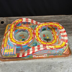 May include: A vintage tin toy roller coaster set featuring a figure-eight track with red and white striped supports and yellow rails. The base includes a Coney Island ticket booth graphic and two small colorful toy cars are placed on the surface.