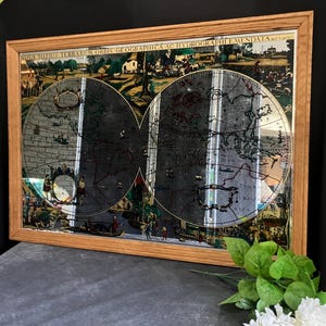 May include: A framed antique world map with a light brown wooden frame. The map features detailed illustrations and text, with a focus on the continents and oceans. The map is displayed on a dark surface with a bouquet of white flowers and green leaves in the foreground.