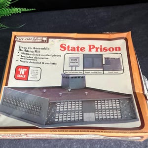 May include: A box for a model building kit of a state prison. The box shows a photo of the assembled model, which is gray and has a guard tower. The box also has the words "State Prison" and "The "Un"welcome Sign" printed on it.