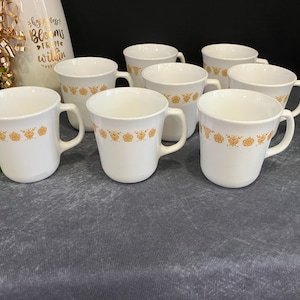 Peut inclure: Ensemble de huit tasses en céramique blanche ornées d'un motif floral jaune et orange. Chaque tasse est dotée d'une anse et d'une forme classique, parfaites pour le café ou le thé.