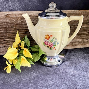 Puede incluir: Una tetera vintage de color crema con un diseño floral, una base plateada y una tapa azul oscuro. La tetera tiene un pico y un asa curvos. Flores amarillas y hojas verdes están a la izquierda.