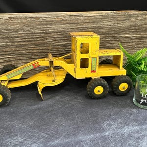 May include: Yellow toy construction grader with the words "Power Grader" and "Luman" on the side. The toy is sitting on a wooden surface with a green plant and a glass jar with the words "Let Love Grow" on it.