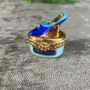 May include: A small oval trinket box with a light blue base and a gold metal rim featuring a floral clasp. The lid is topped with a miniature high-heeled shoe in shades of blue and gold. The word BERMUDA is printed in blue on the front of the box.