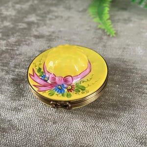 Op de afbeelding: Een klein, rond, geel snuisterijendoosje in de vorm van een zonnehoed. Het deksel is voorzien van een geschilderd roze lint met een strik en kleine blauwe en rode bloemen. De basis is van goudkleurig metaal met een zichtbaar scharnier en sluiting.