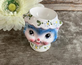 Könnte beinhalten: Ein Eierbecher aus Keramik in Form eines Katzenkopfes mit blauem Fell, großen dunklen Augen und einer rosa Nase. Die Katze trägt einen weißen, gerüschten Kragen, der mit kleinen blauen und gelben Blumen sowie grünen Blättern bemalt ist.