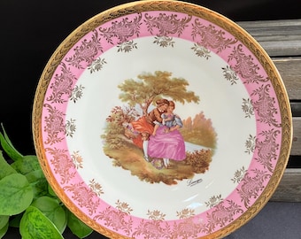 May include: A decorative porcelain plate with a pink and gold border. The center features a romantic scene of a couple under a tree. The plate has a gold rim and is displayed on a clear stand. The plate is a vintage item.