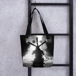 Puede incluir: Bolso tote en blanco y negro con la silueta de una figura con un tocado en forma de cruz frente a una hoguera. El bolso tiene asas negras y está colgado de una escalera de metal negro.