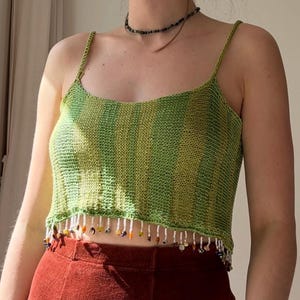 Könnte beinhalten: Ein grün-gelb gestreiftes Strickoberteil mit Spaghettiträgern. Der untere Saum ist mit einem Fransenbesatz aus kleinen, bunten Perlen verziert. Dazu wird ein rostfarbener Rock getragen.