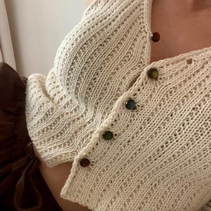Könnte beinhalten: Eine ärmellose Strickweste in Cremefarben mit vertikaler Rippenstruktur und Knopfleiste auf der Vorderseite. Die Weste ist mit fünf kleinen, runden, mehrfarbigen Knöpfen in den Farben Rot, Gelb, Blau, Grün und Braun verziert.