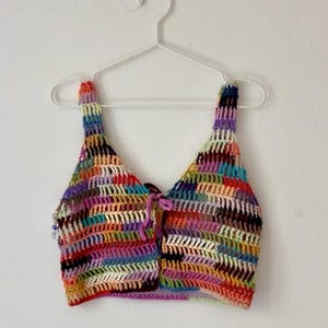 Op de afbeelding: Een kleurrijke, handgemaakte gehaakte top met een V-hals. De top heeft een levendige mix van kleuren, waaronder rood, blauw, geel en paars, waardoor een patchwork-effect ontstaat. Het heeft dunne bandjes en een striksluiting aan de voorkant, hangend aan een witte hanger.