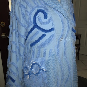 May include: Light blue robe with intricate detailing. The robe features a textured design with darker blue embroidered accents and a shawl collar. The robe has a pocket and decorative buttons. The overall design is artistic and unique.