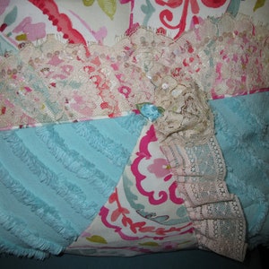 May include: A decorative pillow with a patchwork design featuring a blue, pink, and white floral pattern. The pillow is trimmed with white lace and has a fluffy texture.