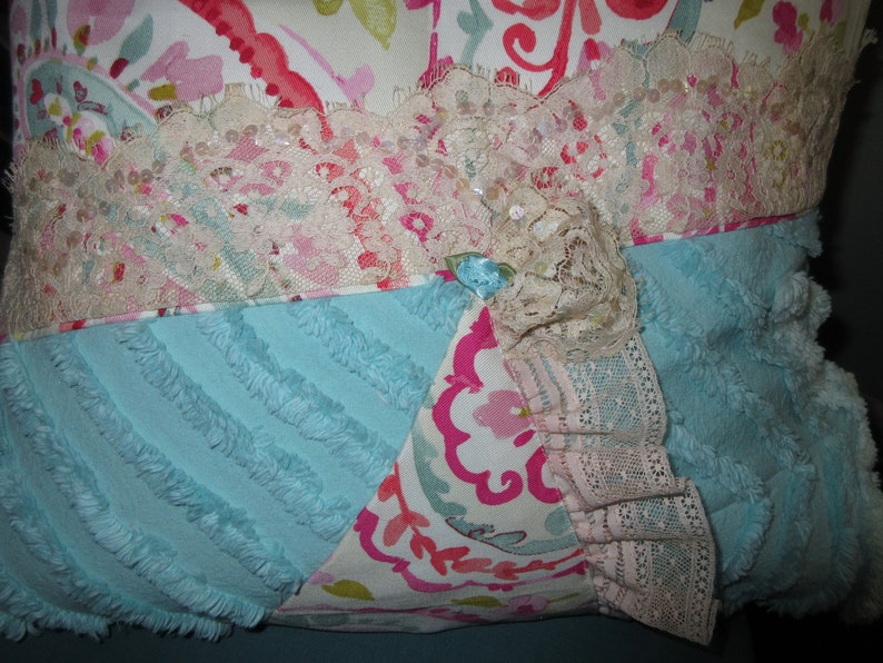 May include: A decorative pillow with a patchwork design featuring a blue, pink, and white floral pattern. The pillow is trimmed with white lace and has a fluffy texture.