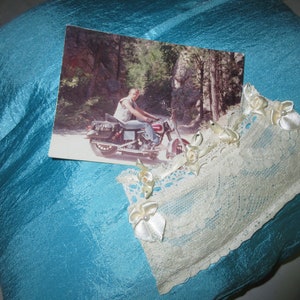 May include: A blue satin pillow with a white lace trim and a photo of a person riding a motorcycle in a wooded area.