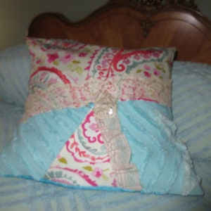 May include: A decorative pillow with a blue, pink, and white paisley pattern. The pillow has a blue and white fluffy texture on one side and a paisley pattern on the other side. The pillow has a large, white, decorative button in the center.