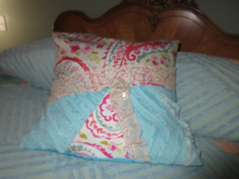 May include: A decorative pillow with a blue, pink, and white paisley pattern. The pillow has a blue and white fluffy texture on one side and a paisley pattern on the other side. The pillow has a large, white, decorative button in the center.