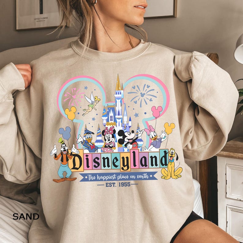 May include: Sand-colored sweatshirt featuring a Disneyland graphic. The design includes the Disneyland logo, a castle, fireworks, and various Disney characters like Mickey and Minnie Mouse. The text reads "Disneyland" and "The happiest place on earth - EST. 1955."