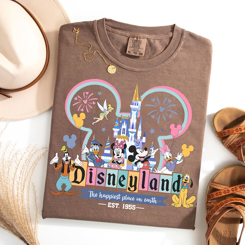 May include: Brown t-shirt with a Disneyland graphic featuring Mickey Mouse, Minnie Mouse, Donald Duck, and other characters. The shirt has the text "Disneyland" and "The happiest place on earth - EST. 1955-". A gold necklace, a hat, and sandals are also in the image.