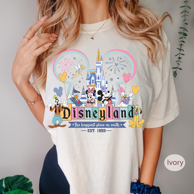 May include: Ivory-colored t-shirt featuring a colorful Disneyland graphic. The design includes the Disneyland logo, iconic characters like Mickey and Minnie Mouse, and a castle. The text "The happiest place on earth - EST. 1955" is also present.