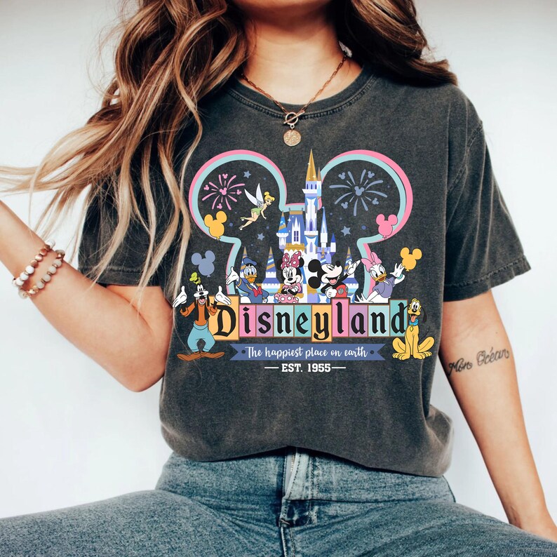 May include: Dark gray t-shirt featuring a colorful Disneyland graphic. The design includes the Disneyland logo, iconic characters like Mickey and Minnie Mouse, and the phrase "The happiest place on earth." The shirt is dated EST. 1955.