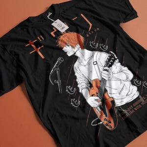Può includere: T-shirt nero con un'illustrazione in stile anime di una persona con i capelli arancioni che suona la chitarra e canta in un microfono. La maglietta ha testo giapponese e la parola "given". La maglietta ha anche il testo "CAN'T SAY GOODBYE, I'M DRIFTING WITH YOU".