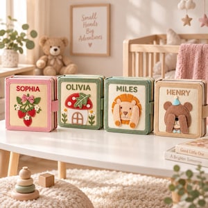 May include: Four square felt books with personalised names and appliqués. The first is pink with strawberries and 'SOPHIA', the second is green with a mushroom and 'OLIVIA', the third is green with a lion and 'MILES', and the fourth is tan with a bear and 'HENRY'.