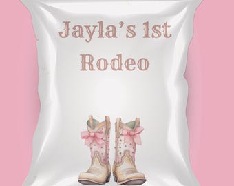 以下が含まれることがあります： 茶色のフォントで「Jayla's 1st Rodeo」というテキストが入った白いパッケージ。 パッケージには、ピンクのリボンが付いた、薄いタンとピンクのカウボーイブーツのペアが描かれています。 背景は無地のピンク色です。