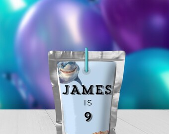 May include: A silver pouch with a light blue label featuring a cartoon shark and the text "JAMES IS 9". The background includes purple and teal balloons, suggesting a birthday theme.